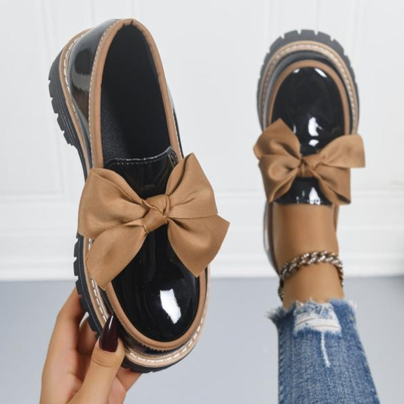 Black patent leather platform loafers with tan bow accent — hand-held and on-foot view
