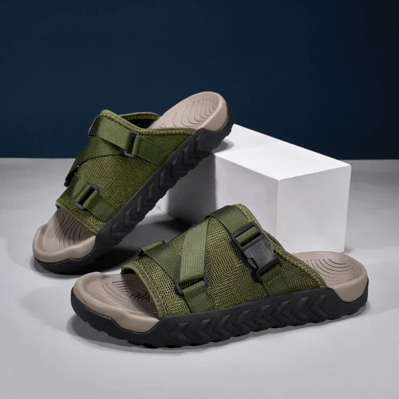 Olive green slip-on sandals with velcro straps and beige footbed on white pedestal — three-quarter front view