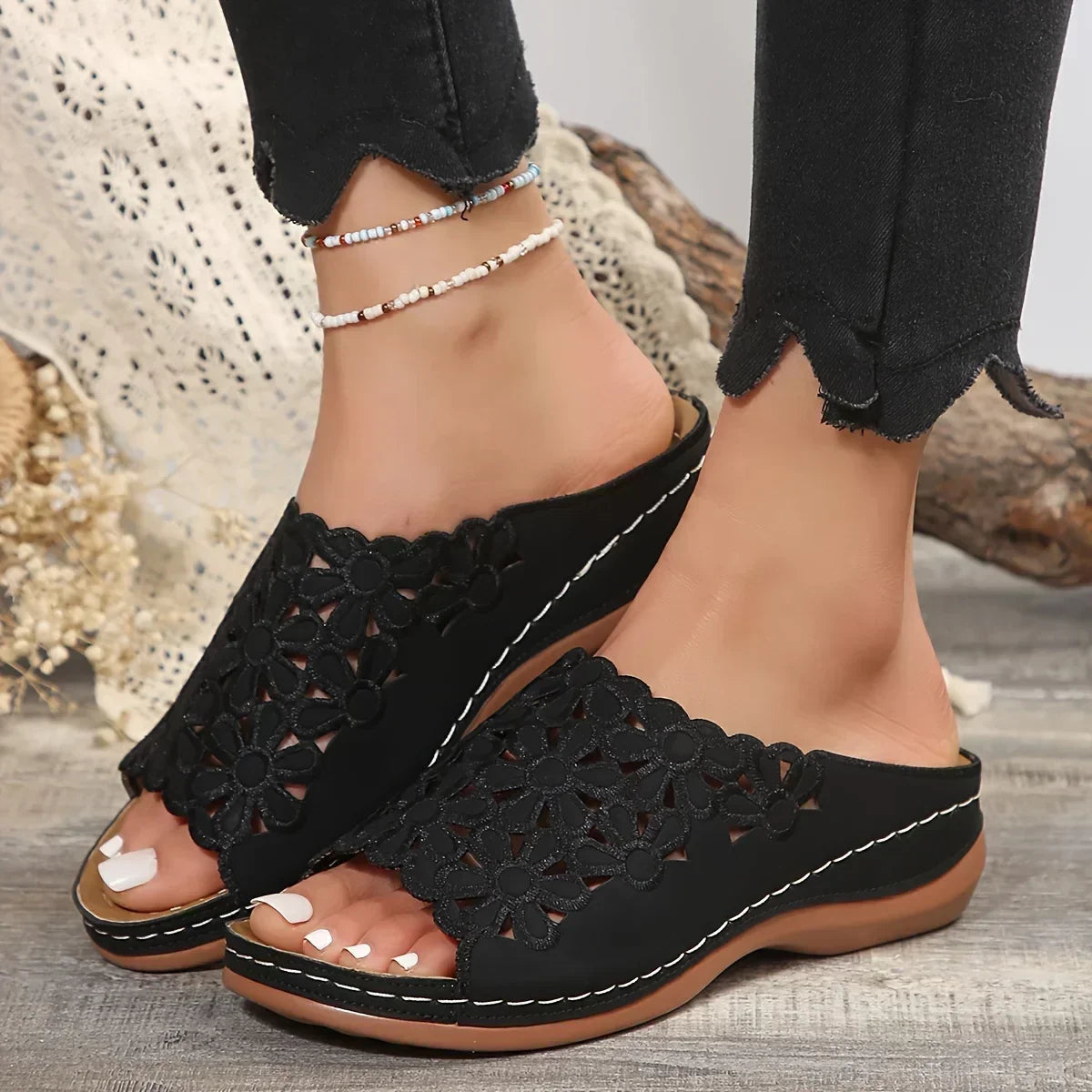 Black floral cutout flat sandals with white stitching on feet wearing black jeans — front angled view